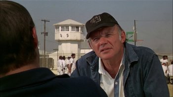 Movie still from “The Longest Yard” (1974), directed by Robert Aldrich – An older man wearing a hat and a jean jacket in front of a prison; Close Up shot, Over the shoulder angle
