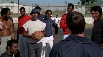 Movie still from “The Longest Yard” (1974), directed by Robert Aldrich – A group of men standing on top of a basketball court; Medium shot, Low angle