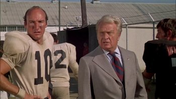 Movie still from “The Longest Yard” (1974), directed by Robert Aldrich – A man in a suit and tie standing next to another man in a football uniform; Medium shot, Over the shoulder angle