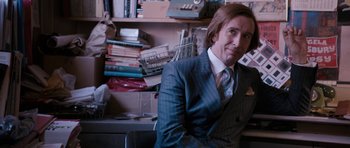 Movie still from “The Look of Love” (2013), directed by Michael Winterbottom – A man in a suit and tie in front of a cluttered desk; Medium shot, Over the shoulder angle