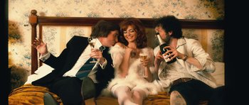 Movie still from “The Look of Love” (2013), directed by Michael Winterbottom – A group of people sitting on a bed drinking champagne; Medium shot, High angle