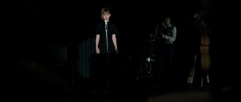 Movie still from “The Look of Love” (2013), directed by Michael Winterbottom – A person standing on a stage with a microphone; Medium shot, Over the shoulder angle