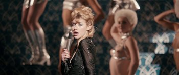 Movie still from “The Look of Love” (2013), directed by Michael Winterbottom – A woman holding a microphone in front of a group of women; Medium shot, Over the shoulder angle