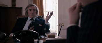 Movie still from “The Look of Love” (2013), directed by Michael Winterbottom – A man sitting at a table holding a cigarette in his hand; Medium shot, Over the shoulder angle