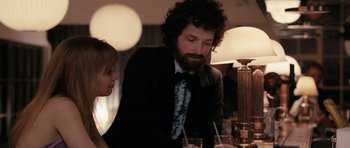 Movie still from “The Look of Love” (2013), directed by Michael Winterbottom – A man in a black suit and a woman in a black suit are sitting at a dinner table; Medium shot, Over the shoulder angle