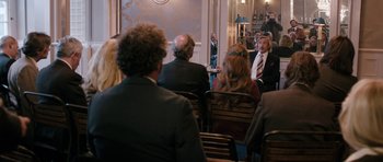 Movie still from “The Look of Love” (2013), directed by Michael Winterbottom – A group of people sitting in chairs in a room; Wide shot, Over the shoulder angle