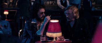 Movie still from “The Look of Love” (2013), directed by Michael Winterbottom – A man sitting at a table next to another man; Medium shot, Over the shoulder angle