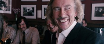 Movie still from “The Look of Love” (2013), directed by Michael Winterbottom – A man with long blonde hair and a mustache smiling for the camera; Close Up shot, Over the shoulder angle