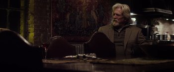 Movie still from “The Lords of Salem” (2012), directed by Rob Zombie – An older man sitting at a table in front of a painting; Medium shot, Over the shoulder angle