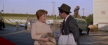 Movie still from “The Lost World” (1960), directed by Irwin Allen – An older woman holding a small dog while a man in a suit and hat looks on; Medium shot, Over the shoulder angle