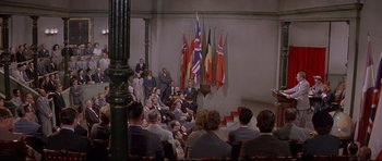 Movie still from “The Lost World” (1960), directed by Irwin Allen – A group of people sitting in front of flags; Wide shot, High angle