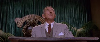 Movie still from “The Lost World” (1960), directed by Irwin Allen – An older man wearing a suit and tie in front of a microphone; Close Up shot, Low angle