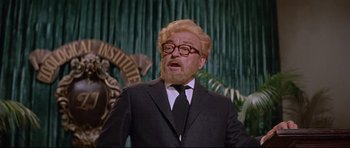 Movie still from “The Lost World” (1960), directed by Irwin Allen – An older man in a suit and tie in front of a green curtain; Medium shot, Low angle