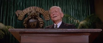 Movie still from “The Lost World” (1960), directed by Irwin Allen – An older man in a suit and tie sitting in front of a microphone; Close Up shot, Low angle