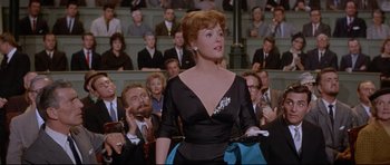 Movie still from “The Lost World” (1960), directed by Irwin Allen – A woman in a black dress standing in front of an audience; Medium shot, Over the shoulder angle