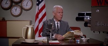Movie still from “The Lost World” (1960), directed by Irwin Allen – An older man sitting at a desk in front of an american flag; Medium shot, Low angle