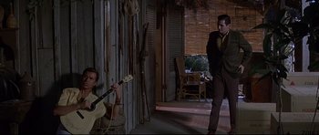 Movie still from “The Lost World” (1960), directed by Irwin Allen – A man in a suit and tie playing a guitar in front of another man in a suit and tie in front of an open door; Wide shot, Over the shoulder angle