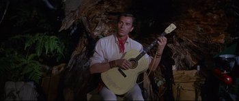 Movie still from “The Lost World” (1960), directed by Irwin Allen – A man sitting down holding a guitar in front of a tree trunk; Medium shot, Low angle