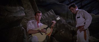 Movie still from “The Lost World” (1960), directed by Irwin Allen – A man sitting on a rock holding a guitar in front of a rock wall; Medium shot, Low angle