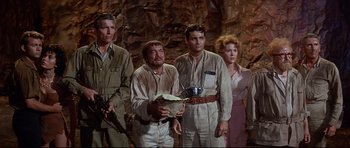 Movie still from “The Lost World” (1960), directed by Irwin Allen – A group of people standing next to each other in front of a rock wall; Medium shot, High angle