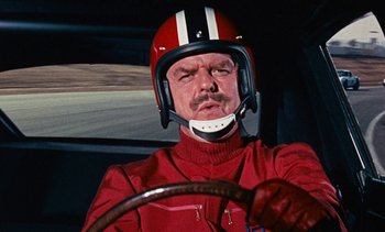 Movie still from “The Love Bug” (1969), directed by Robert Stevenson – A man in a red racing suit driving a race car; Close Up shot, Low angle