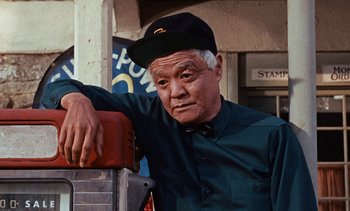 Movie still from “The Love Bug” (1969), directed by Robert Stevenson – An older man wearing a hat leaning on a stack of books; Medium shot, Low angle
