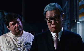 Movie still from “The Love Bug” (1969), directed by Robert Stevenson – A man in a suit and tie standing next to another man; Medium shot, Over the shoulder angle