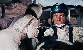 Movie still from “The Love Bug” (1969), directed by Robert Stevenson – A man wearing a helmet driving a car while another man looks on; Close Up shot, Over the shoulder angle