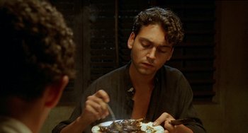 Movie still from “The Lover” (1992), directed by Jean-Jacques Annaud – A man sitting at a table with a plate of food in front of him; Close Up shot, Over the shoulder angle
