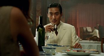 Movie still from “The Lover” (1992), directed by Jean-Jacques Annaud – A man sitting at a table with a glass of wine; Medium shot, Over the shoulder angle