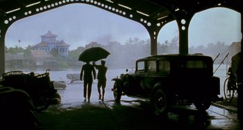 Movie still from “The Lover” (1992), directed by Jean-Jacques Annaud – Two men holding an umbrella under an awning near an antique car; Wide shot, High angle