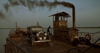 Movie still from “The Lover” (1992), directed by Jean-Jacques Annaud – An old car is parked next to a building with smoke billowing out of it; Extreme Wide shot, High angle