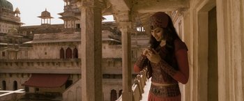 Movie still from “The Lovers” (2015), directed by Roland Joffé – A beautiful woman in a red and brown outfit looking at her cell phone; Medium shot, Over the shoulder angle