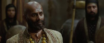 Movie still from “The Lovers” (2015), directed by Roland Joffé – A bald man with a beard and a necklace of beads; Close Up shot, Over the shoulder angle