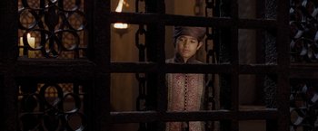 Movie still from “The Lovers” (2015), directed by Roland Joffé – A young boy wearing a hat standing behind a chain link fence; Medium shot, Over the shoulder angle