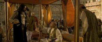 Movie still from “The Lovers” (2015), directed by Roland Joffé – A man sitting on a chair in a room with other people; Wide shot, Over the shoulder angle