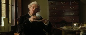 Movie still from “The Lovers” (2015), directed by Roland Joffé – A man in a wig holding a cup in his hand; Medium shot, High angle