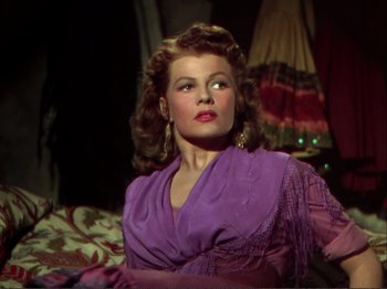 Movie still from “The Loves of Carmen” (1948), directed by Charles Vidor – A woman in a purple dress sitting on top of a couch; Medium shot, Low angle