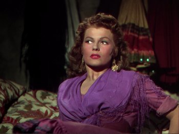 Movie still from “The Loves of Carmen” (1948), directed by Charles Vidor – A woman in a purple dress sitting on a bed; Medium shot, Over the shoulder angle