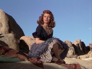 Movie still from “The Loves of Carmen” (1948), directed by Charles Vidor – A woman sitting on top of a large rock; Medium shot, Low angle