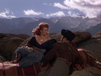 Movie still from “The Loves of Carmen” (1948), directed by Charles Vidor – A woman sitting on top of a rock next to a man; Wide shot, Low angle