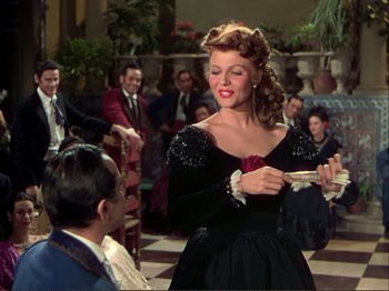 Movie still from “The Loves of Carmen” (1948), directed by Charles Vidor – A woman in a long black dress holding a pen in her hand; Medium shot, Over the shoulder angle