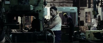 Movie still from “The Machinist” (2004), directed by Brad Anderson – A man working on a machine in an industrial setting; Medium shot, Low angle