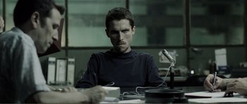 Movie still from “The Machinist” (2004), directed by Brad Anderson – A man sitting in front of a microphone in a room; Medium shot, Over the shoulder angle