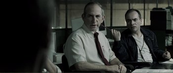 Movie still from “The Machinist” (2004), directed by Brad Anderson – An older man sitting in an office chair; Medium shot, Over the shoulder angle