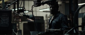 Movie still from “The Machinist” (2004), directed by Brad Anderson – A man working in an industrial setting with a lamp; Medium shot, Over the shoulder angle
