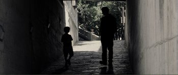Movie still from “The Machinist” (2004), directed by Brad Anderson – A man and a boy standing on a brick walkway; Wide shot, Over the shoulder angle