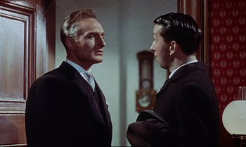 Movie still from “The Man Who Could Cheat Death” (1959), directed by Terence Fisher – Two men in suits talking to each other in front of a wall clock; Medium shot, Over the shoulder angle