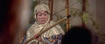 Movie still from “The Man Who Fell to Earth” (1976), directed by Nicolas Roeg – A woman in a gold outfit holding a wooden stick; Close Up shot, High angle