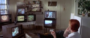 Movie still from “The Man Who Fell to Earth” (1976), directed by Nicolas Roeg – A man sitting in front of two televison sets holding remotes; Medium shot, High angle